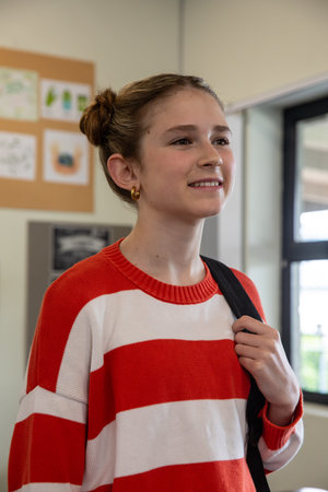 School-age girl in red sweater standing in classroom holding backpack by bulletin board and window....
