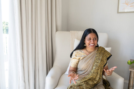 Senior Indian Asian woman wearing sari sitting at home on armchair with artwork and potted plant. Elegant, sophisticated, tranquil, refined, domestic, stylish, contemplativeの写真素材