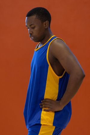 African American man standing in gym by orange wall, wearing blue yellow basketball jersey, shorts....
