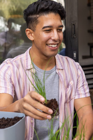 Man potting green shoots with clump of soil into grey planter on patio with wooden shelf. Gardening,...