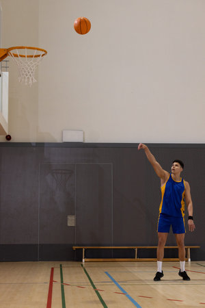Male basketball player in blue-yellow jersey shooting ball midair in gym hoop visible, copy space....