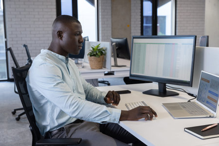 African American man working on spreadsheet on external monitor at modern office, copy space. Data,...