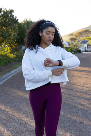 Woman checking wrist smartwatch while standing on paved residential street, wearing hoodie....