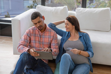 Diverse expecting couple using tablet sitting on floor at home by white couch holding credit card....