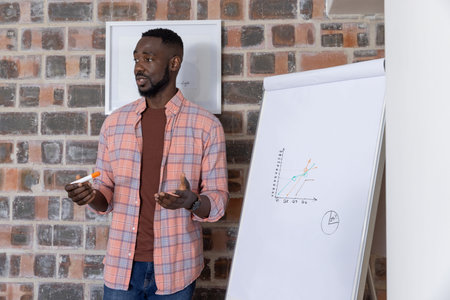 African American man standing beside flip chart explaining graph in office with artwork, copy space....