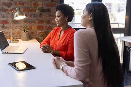 Diverse female colleagues sitting at white desk in modern office area with laptop and tablet. Corporate, teamwork, collaboration, modern, professional, workspace, urbanの写真素材