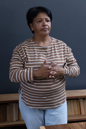 Senior Indian woman standing in room by dark grey wall with shelf of books, clasping hands....