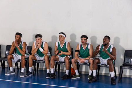 Diverse male basketball teammates in green jerseys and shorts sitting in gym holding basketball....