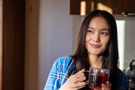 Asian woman holding glass mug of red tea in kitchen with stainless steel kettle, copy space. Candid,...