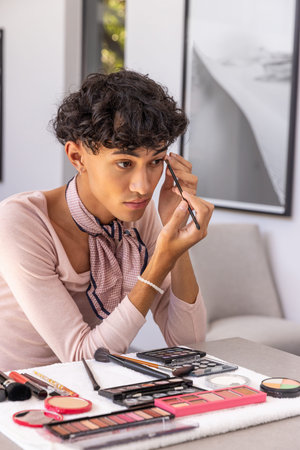 Man wearing pink top and scarf applying eyeliner with brush at home with eyeshadow palettes. Beauty, cosmetics, grooming, artistry, minimalist, serene, personal careの写真素材