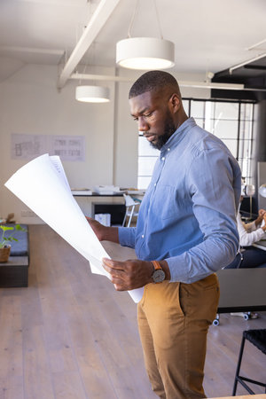 African American man standing and examining blueprints at office desk in workspace, copy space. Professional, workspace, modern, architecture, design, interior, planningの写真素材