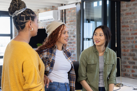 Diverse female coworkers discussing ideas at desk with lamp and potted plant in open plan office. Collaboration, teamwork, brainstorming, modern, urban, professional, casualの写真素材