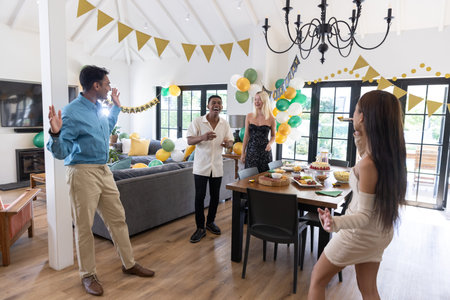 Diverse friends gathering around dining table laughing and pointing at tiered cake in living room. Celebration, friendship, joyful, festive, stylish, decor, gathering