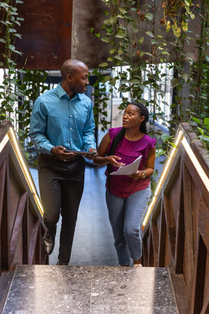 African American coworkers climbing office stairs under vines and LED railings with tablet, papers....
