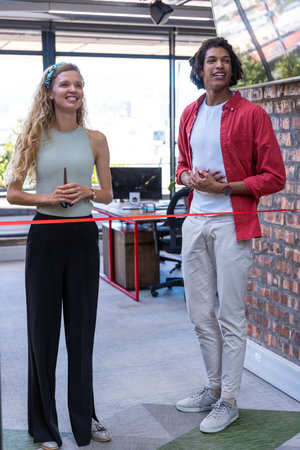 Diverse coworkers cutting red ribbon with scissors at modern open-plan office near desk....