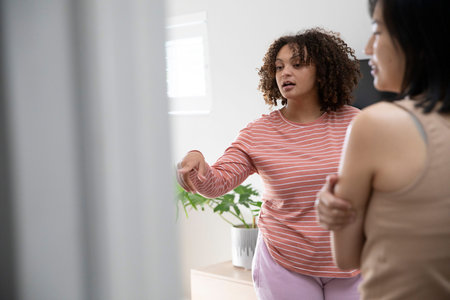 Diverse female friends talking and gesturing in bright room near white dresser and potted plant....