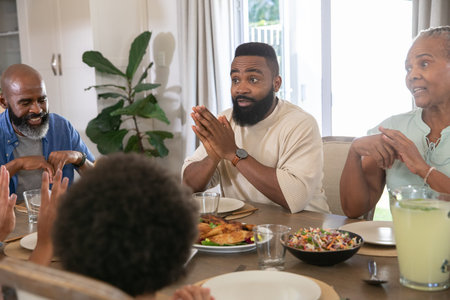 Diverse family bowing heads and clasping hands in home dining room around wooden table before meal....