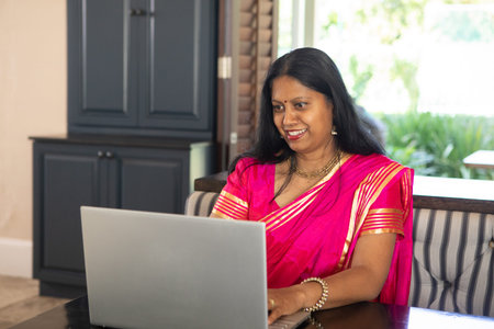 Mature Indian woman wearing pink saree with gold jewelry working on silver laptop at home office....