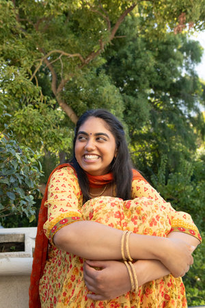 Indian woman hugging knees while smiling on park bench wearing floral kameez and red dupatta....