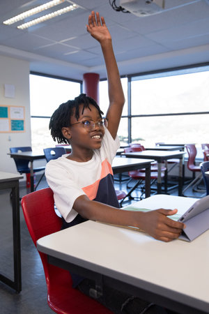 African American boy student raising hand while holding tablet with keyboard at desk in classroom....