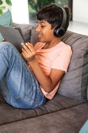 South Asian school-age boy sitting on gray sofa in living room holding tablet wearing headphones....