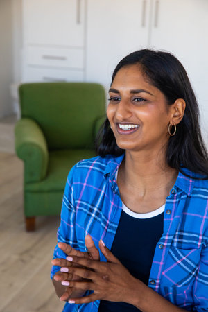 Indian woman sitting in living room, gesturing near green armchair, white cabinets, copy space....