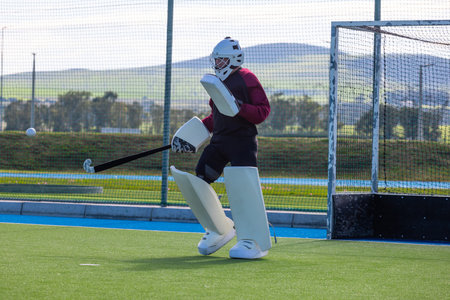 Male field hockey goalkeeper standing in helmet and pads on synthetic turf with ball, copy space. Athletic, action, competitive, sport, outdoor, vibrant, determinationの写真素材