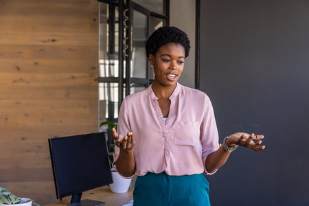 African American woman gesturing and speaking at office desk with wooden wall, laptop and plant....