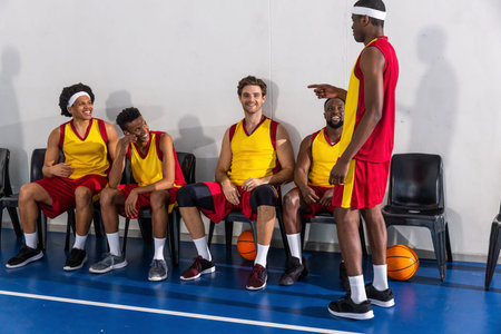 Diverse male basketball teammates sitting on sideline in gym laughing and pointing at basketballs....
