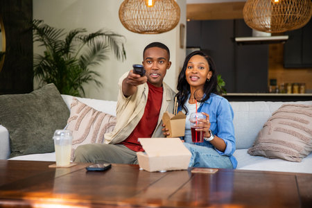 African American couple sitting on sofa holding TV remote and take-out drink in living room. Modern,...