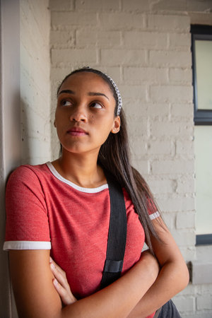 Teenage girl leaning against brick wall in hall wearing headband carrying black shoulder bag strap....