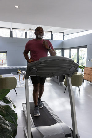Senior African American man wearing sportswear running on treadmill in modern living room. Fitness,...