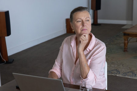 Senior woman sitting at table in quiet room using laptop and holding glass of water. Computer, home,...
