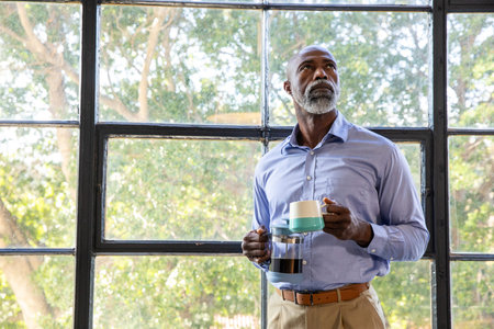 Senior African American man in blue shirt standing by grid window holding carafe, mugs, copy space....