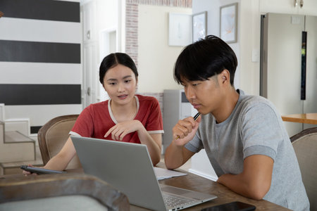 Asian couple at wooden dining table, woman in red shirt holding tablet, pointing at laptop papers....