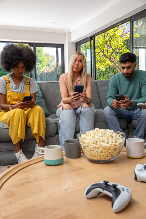Diverse friends using smartphones on sofa in living room near popcorn bowl, one in yellow overalls....