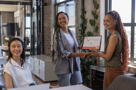 Diverse female coworkers exchanging certificate and shaking hands at office table with laptop....