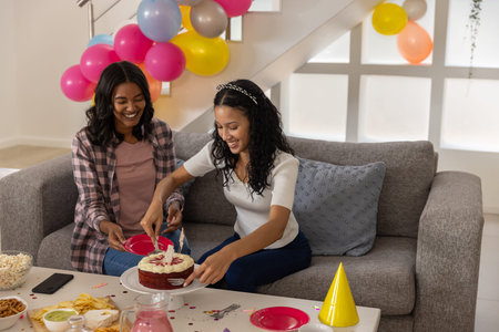 Mom and teenage daughter cutting birthday cake with lit candles in living room with pastel balloons....