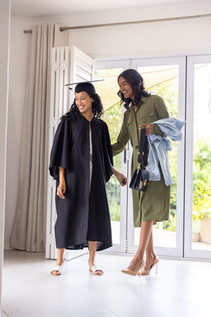 African American mother guiding teenage daughter in home glass doorway holding graduation cap, gown. Celebration, achievement, family, milestone, elegance, outdoor, warmthの写真素材