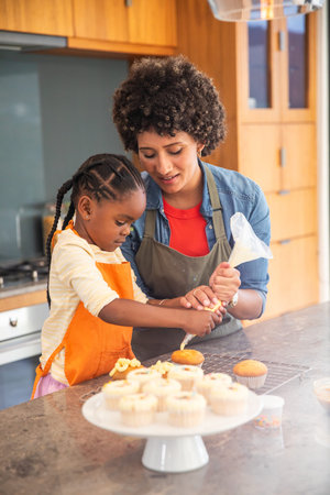 Mother and daughter decorating cupcakes at countertop in home kitchen using frosting piping bag....