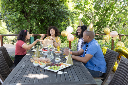 Diverse friends sitting around table on deck sharing gift bag and sampling pastries amid balloons....