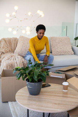 African American woman unpacking boxes on sofa next to coffee cup and smartphone in living room....