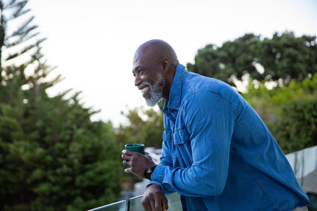 Senior African American man leaning on glass railing with green mug overlooking garden, copy space....