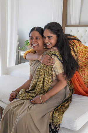 Indian mother and daughter wearing traditional attire embracing and smiling on bed in bedroom....