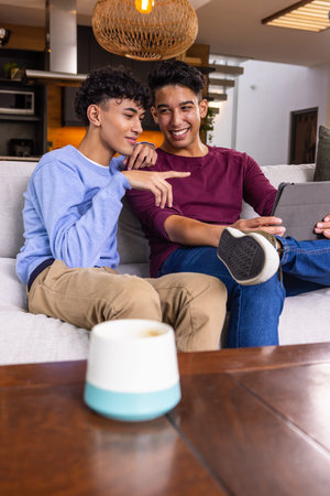 Two male friends sitting on sofa in open-plan living area pointing at tablet. Friendly, casual,...