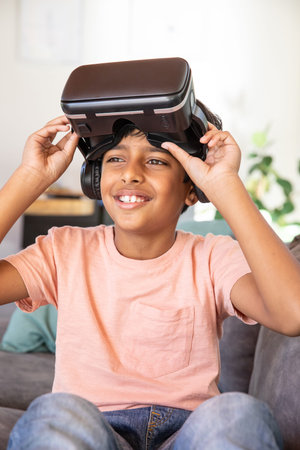 Indian boy sitting on sofa holding VR headset above eyes while wearing headphones in living room....