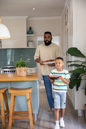African American father and son walking through kitchen carrying bowl and tray past island stovetop. Family, bonding, domestic, cozy, home, nourishment, lifestyleの写真素材