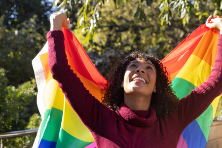 African American woman wearing sweater holding rainbow-striped pride flag in park smiling upward....