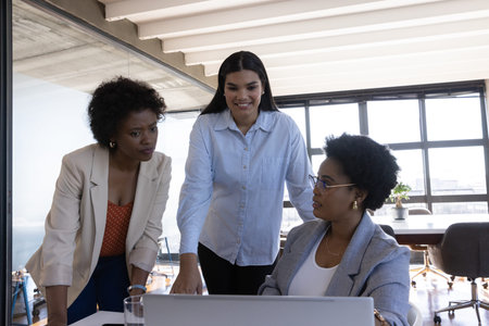 Diverse female coworkers collaborating around laptop on meeting table in conference room....