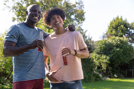 Diverse male friends standing side by side in backyard holding red drink bottles and barbecue tongs....